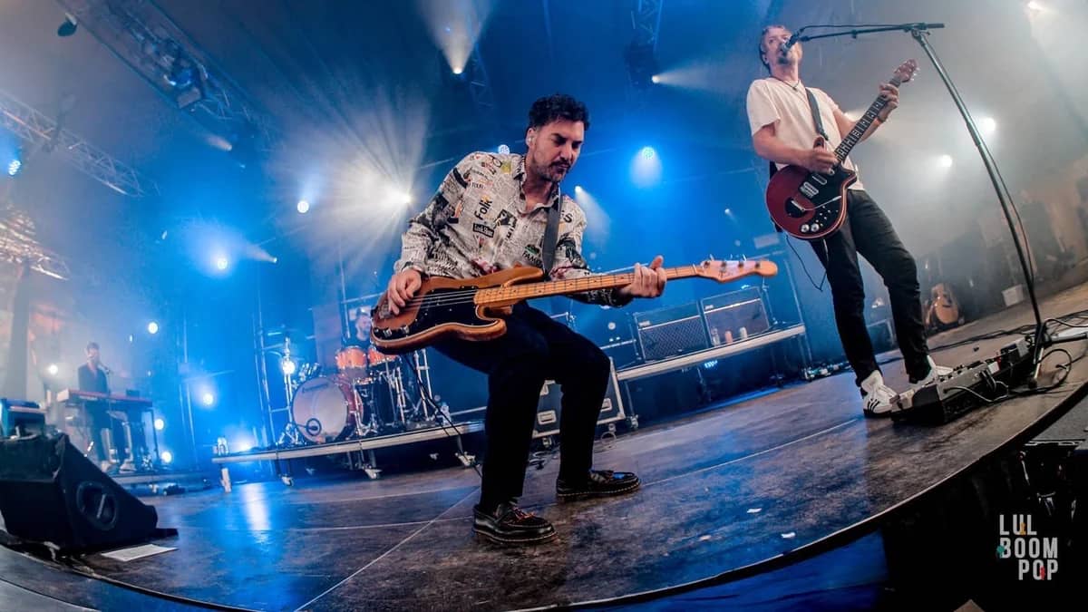 Bassist van The Dutch Queen speelt energiek op het podium met gitarist op de achtergrond, verlicht door blauw podiumlicht op een festival