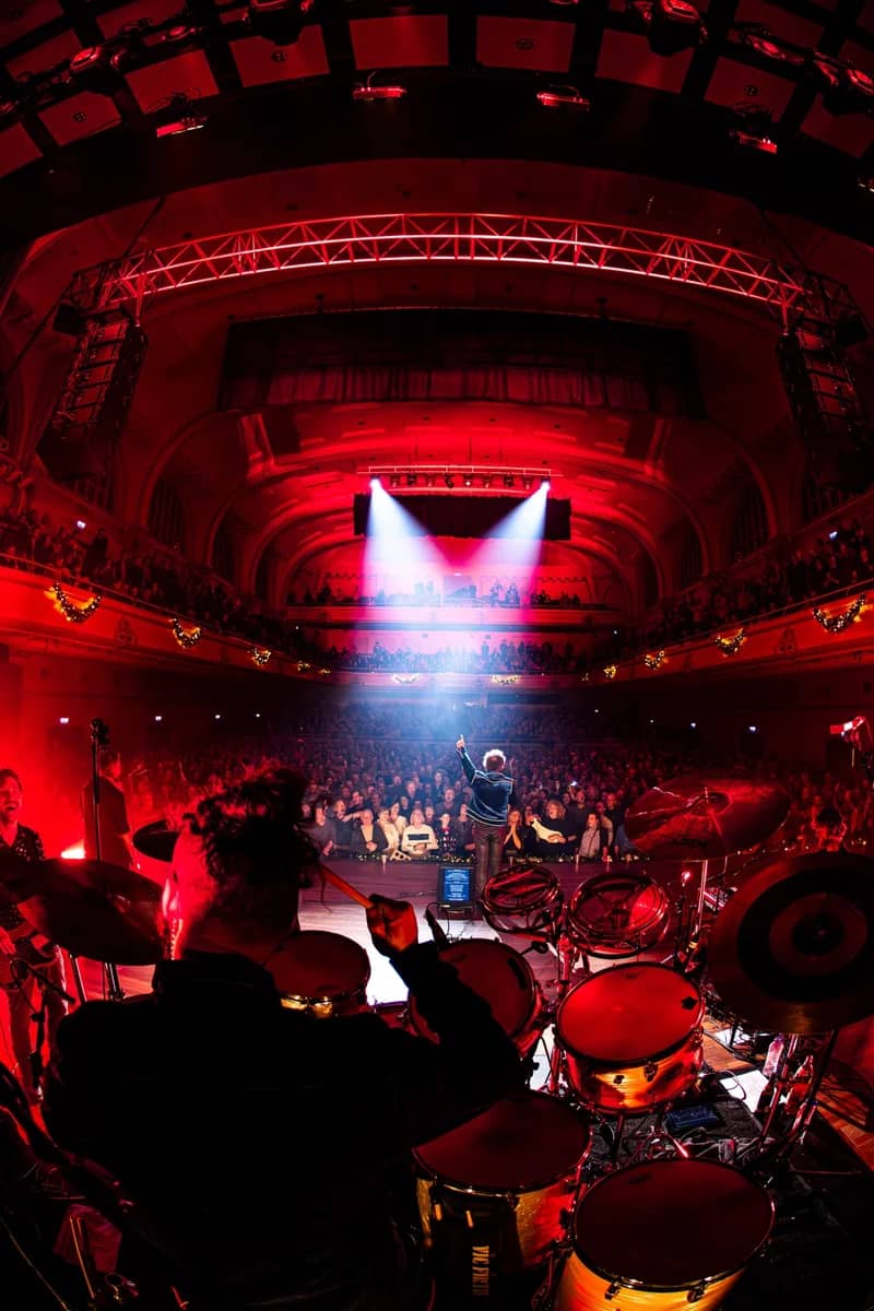 Uitzicht vanaf het drumstel over een uitverkochte theaterzaal in rood licht, zanger met opgeheven arm richting het publiek