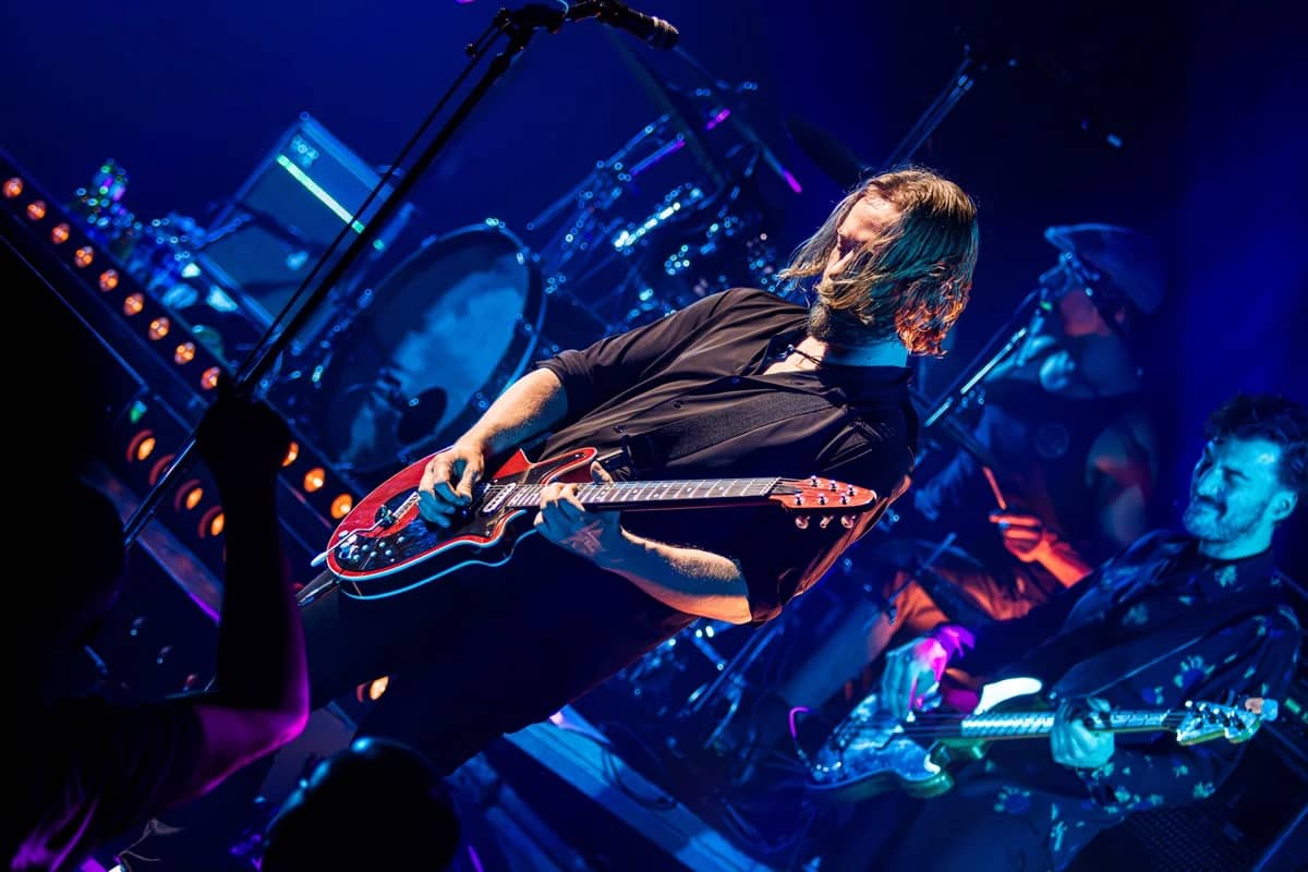 Gitarist van The Dutch Queen speelt een rode gitaar met volle overgave, blauwe podiumverlichting en drummer op de achtergrond
